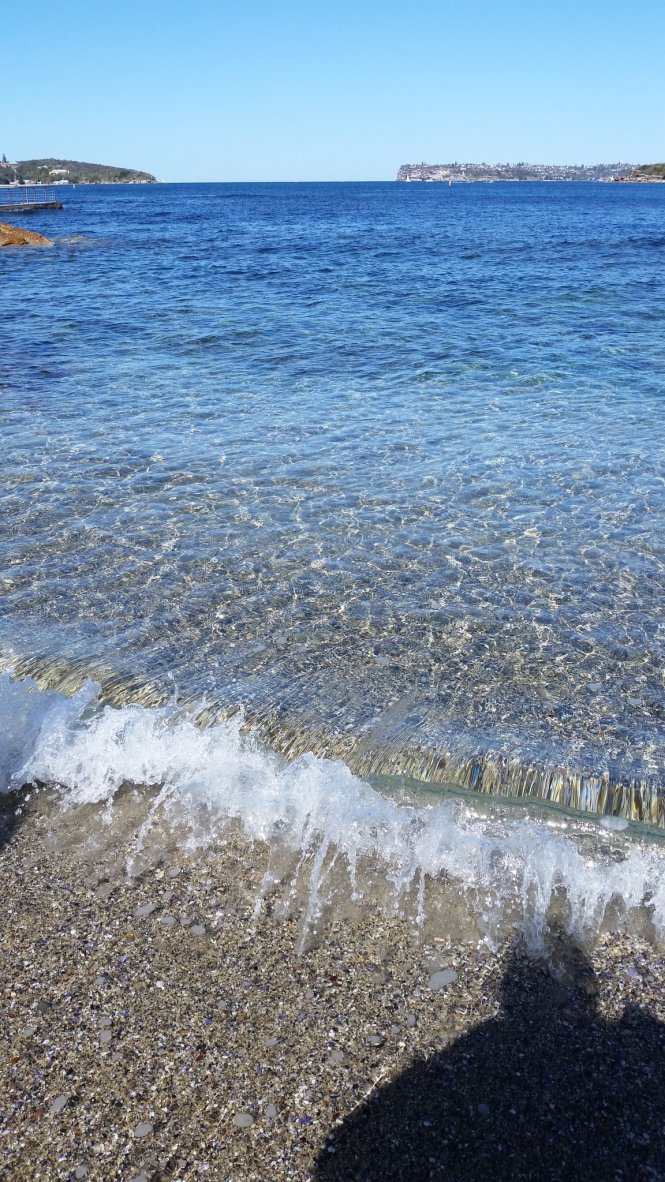 A long shot of a calm body of harbour water with a small wave at the shallows. There is a person's shadow in the corner near the shallow wave
