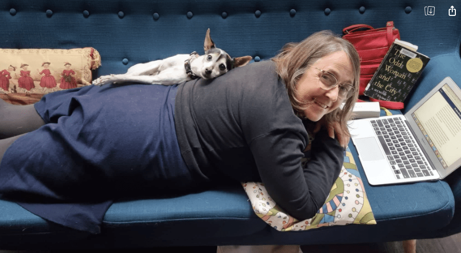 A white dog lying on my back. I have a blue skirt, a black top on and there is a novel next to me. We are on a blue couch.