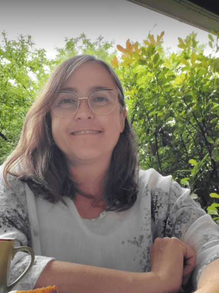 A photograph of myself wearing a white top with blue flowers. Backdrop of green plants and a small demitasse of coffee by my arm.