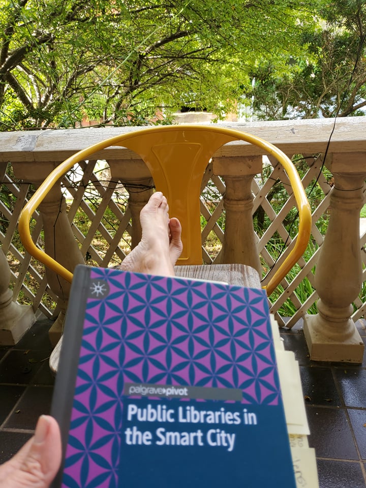 A photograph of the book cover (very boring blue and purple pattern with the book title in white). I am holding the book with my feet on a tolix chair overlooking my front verandah which is framed with greenery from the lovely trees.