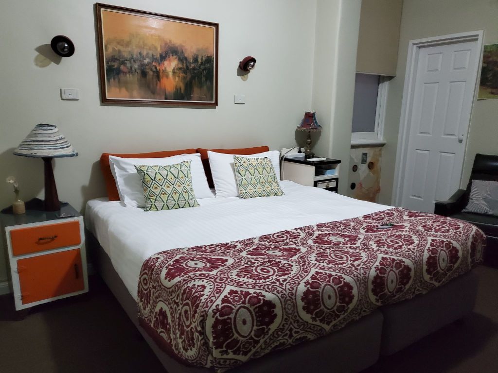 A bed with white linen, red blanket at the foot, orange bedside table and orange paining like a smudge.