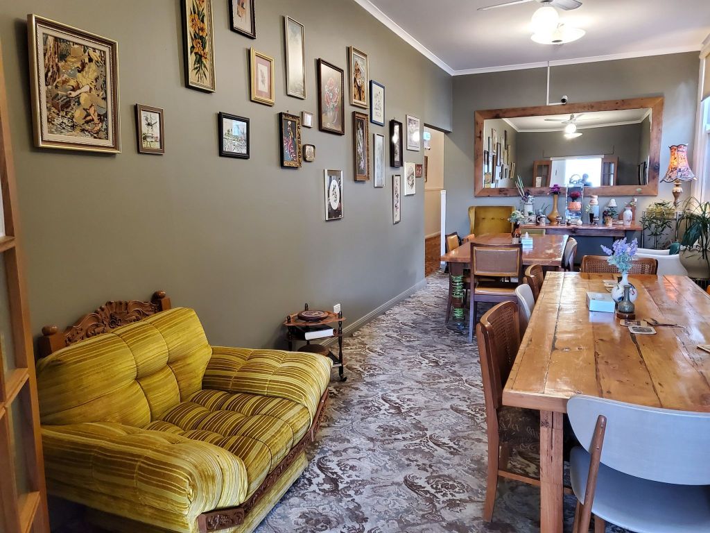 A dining room with two long tables, a mishmash of chairs and lounges, a large mirror and a wall of scattered framed embroideries.