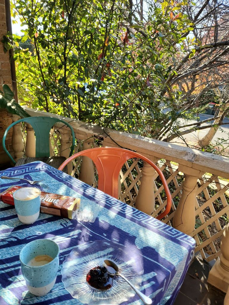 2 coffees, timtams and spoon sweet, on a blue tablecloth with a Greek motif, orange and green verandah chairs. Columns and lattice.
